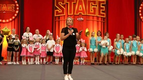 eine junge Frau in schwarzer Sportkleidung steht auf einer rotbeleuchteten Bühne. Im Hintergrund viele Kinder in Kostümen