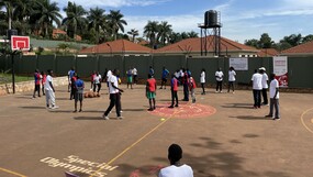Ein Outdoor-Sportplatz mit Basketballkörben in Afrika und ca. 30 jungen Afrikaner*innen in unterschiedlichen Trikots